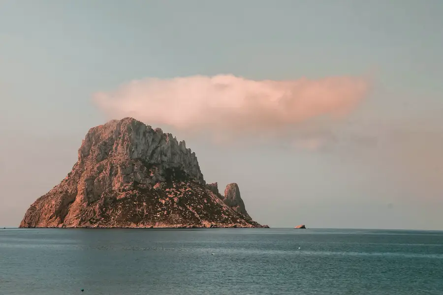 The rock of Es Vedrà rising from the sea under a soft pink cloud.